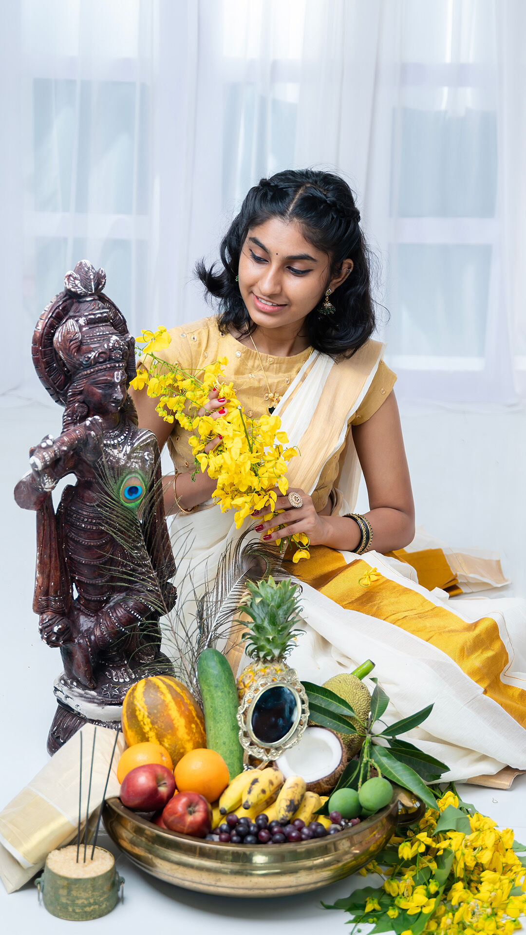 Young girl in traditional Kerala attire offering yellow flowers to a wooden Krishna idol, with a festive brass plate of fruits and mirror arranged for Janmashtami celebration